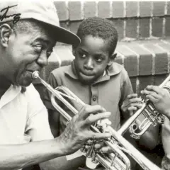 Louis Armstrong enseña a dos niños a tocar la trompeta. Ella Fitzgerald, aunque no está en la imagen, es una leyenda del jazz junto a él.
