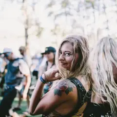 Chica sonriente con tatuajes en un festival de música electrónica, rodeada de gente bailando bajo el sol.