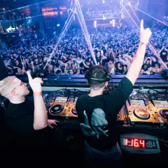 Dos DJs en perfecta alignment, con las manos en alto, animan a una multitud vibrante en una discoteca con luces láser.
