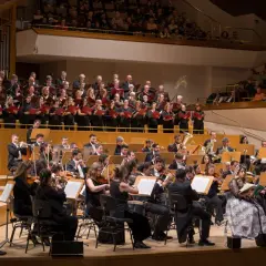 Un coro y orquesta interpretan música en un auditorio. Los cantantes franceses, vestidos de negro, sostienen partituras rojas.