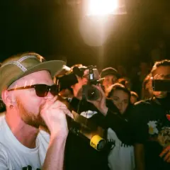 Un rapero con gorra y gafas de sol canta al micrófono, rodeado de público y cámaras. Capturando la energía de los estilos de música urbana.