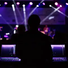 Silueta de técnico de sonido operando una mesa de mezclas en un plano de escenario musical, con luces púrpuras y blancas iluminando la banda al fondo.