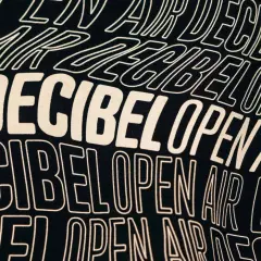 Texto en una camiseta negra con el lema "Decibel Open Air" repetido en letras blancas onduladas, evocando la energía de un festival.
