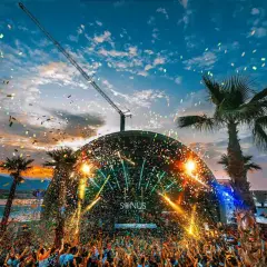 Multitud celebra en el festival croacia techno bajo confeti y luces. Palmeras y grúa completan la escena.