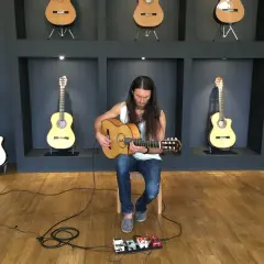 Un guitarrista con barba toca una guitarra acústica sentado frente a pedales de efectos.