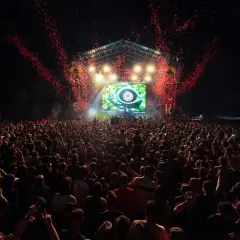 Multitud en un concierto al aire libre, con confeti rojo cayendo sobre el escenario y una pantalla gigante. El **bass** retumba en el parque.
