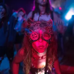 Una persona con una máscara de gato steampunk y gafas de aviador, rodeada de luces de neón y gente bailando. Así es que es la rave.