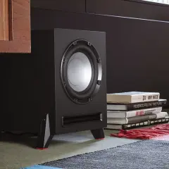 Subwoofer negro con detalles rojos, junto a libros y una alfombra.
