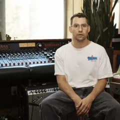 Productor musical con gafas sentado frente a una mesa de mezclas y un teclado en un estudio de grabación.
