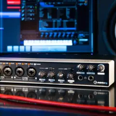 Interfaz de audio Steinberg UR44 con cable rojo conectado, monitor de computadora y altavoz al fondo.