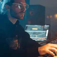 Hombre con gafas y coleta trabajando en un estudio de sonido, creando música electrónica para su blog.