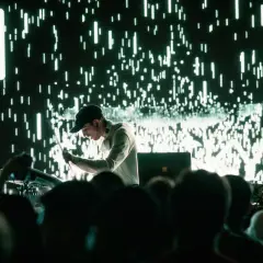 Un DJ techno en acción, con una pantalla de luces verdes y blancas parpadeantes detrás.