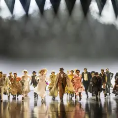 Un grupo diverso de personas corre hacia adelante en un escenario, evocando la energía y el espíritu de "Ragtime, que es".