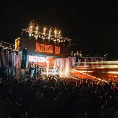 Una multitud celebra en un concierto nocturno, con fuegos artificiales y luces láser iluminando el escenario "AREA 19" y el **nexus fabrik** de contenedores.