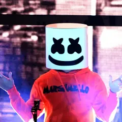Christopher Comstock, conocido como Marshmello, con su icónica máscara blanca y sudadera naranja, saluda al público en un concierto.