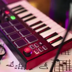 Teclado rojo con pads, botones y teclas blancas y negras. Unas gafas negras y partituras debajo. Es un controlador MIDI para crear música.