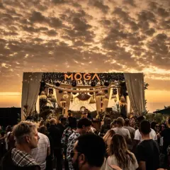 Multitud disfrutando del atardecer en el moga festival, con luces y escenario decorado.