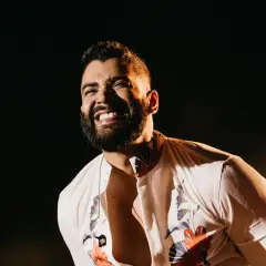 Un cantante brasileño famoso joven ríe con alegría en el escenario, con una camisa blanca estampada y un collar.