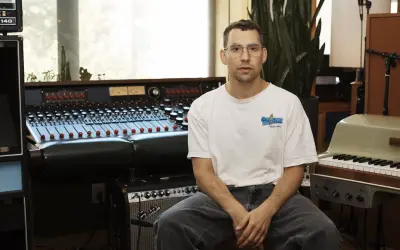 Productor musical con gafas sentado frente a una mesa de mezclas y un teclado en un estudio de grabación.