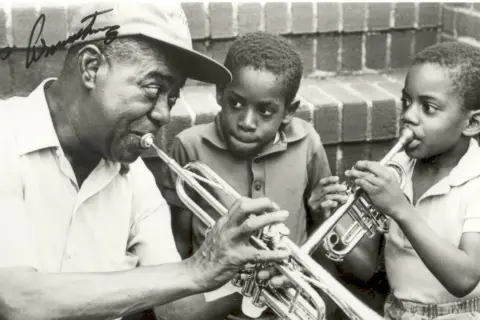 Louis Armstrong enseña a dos niños a tocar la trompeta. Ella Fitzgerald, aunque no está en la imagen, es una leyenda del jazz junto a él.