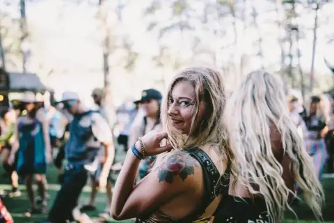Chica sonriente con tatuajes en un festival de música electrónica, rodeada de gente bailando bajo el sol.