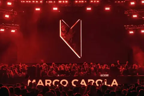 Multitud vibrante en Madrid, luces rojas y el logo de Marco Carola iluminan la noche.