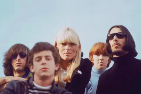 Cinco miembros de The Velvet Underground, pioneros del sonido new wave 80, posan con miradas intensas bajo un cielo azul.