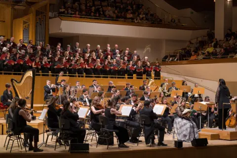 Un coro y orquesta interpretan música en un auditorio. Los cantantes franceses, vestidos de negro, sostienen partituras rojas.