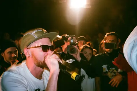 Un rapero con gorra y gafas de sol canta al micrófono, rodeado de público y cámaras. Capturando la energía de los estilos de música urbana.