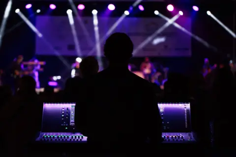 Silueta de técnico de sonido operando una mesa de mezclas en un plano de escenario musical, con luces púrpuras y blancas iluminando la banda al fondo.