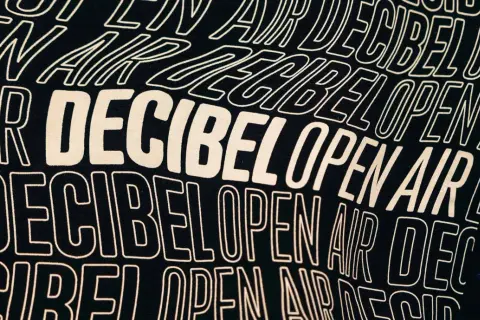 Texto en una camiseta negra con el lema "Decibel Open Air" repetido en letras blancas onduladas, evocando la energía de un festival.