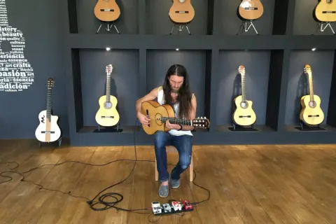 Un guitarrista con barba toca una guitarra acústica sentado frente a pedales de efectos.