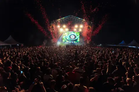 Multitud en un concierto al aire libre, con confeti rojo cayendo sobre el escenario y una pantalla gigante. El **bass** retumba en el parque.