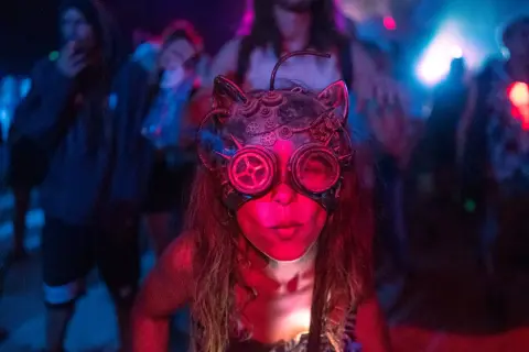 Una persona con una máscara de gato steampunk y gafas de aviador, rodeada de luces de neón y gente bailando. Así es que es la rave.