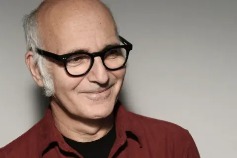Retrato de Ludovico Einaudi, músico italiano. Su edad se refleja en su mirada serena y su cabello canoso. Viste una camisa roja.