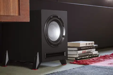 Subwoofer negro con detalles rojos, junto a libros y una alfombra.