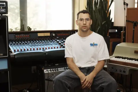 Productor musical con gafas sentado frente a una mesa de mezclas y un teclado en un estudio de grabación.