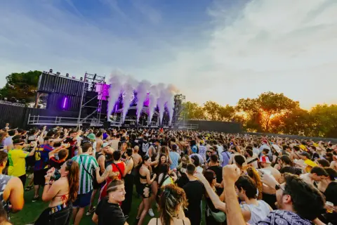 Multitud bailando en un festival al atardecer, con humo y luces moradas saliendo del escenario. ¡Un ambiente genial para los parkineos de donde es!