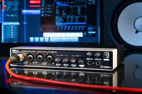 Interfaz de audio Steinberg UR44 con cable rojo conectado, monitor de computadora y altavoz al fondo.
