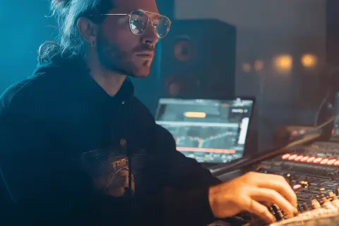 Hombre con gafas y coleta trabajando en un estudio de sonido, creando música electrónica para su blog.