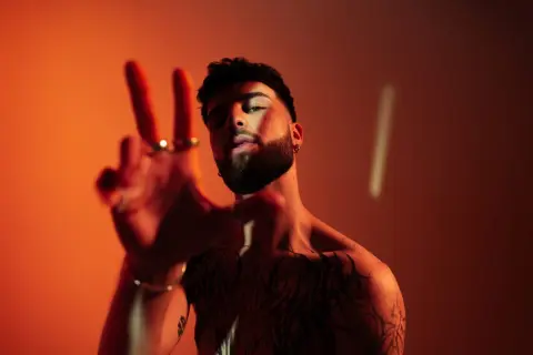 Joven con tatuajes y barba hace el signo de la paz. ¿Oguz DJ de dónde es?