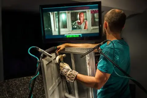 Un técnico de sonido crea efectos de **foley que es** el arte de añadir sonidos a una película, golpeando una ventana con un martillo.