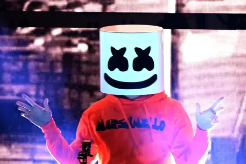 Christopher Comstock, conocido como Marshmello, con su icónica máscara blanca y sudadera naranja, saluda al público en un concierto.