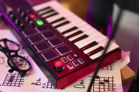 Teclado rojo con pads, botones y teclas blancas y negras. Unas gafas negras y partituras debajo. Es un controlador MIDI para crear música.