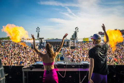 Dos DJs en un escenario lanzan fuego al aire ante una multitud masiva en uno de los festivales techno de Europa.