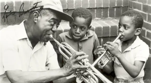 Louis Armstrong enseña a dos niños a tocar la trompeta. Ella Fitzgerald, aunque no está en la imagen, es una leyenda del jazz junto a él.
