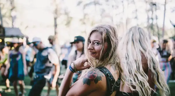 Chica sonriente con tatuajes en un festival de música electrónica, rodeada de gente bailando bajo el sol.