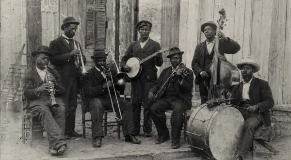 Grupo de músicos tocando jazz años 50: clarinete, trombón, banjo, violín, contrabajo y batería.