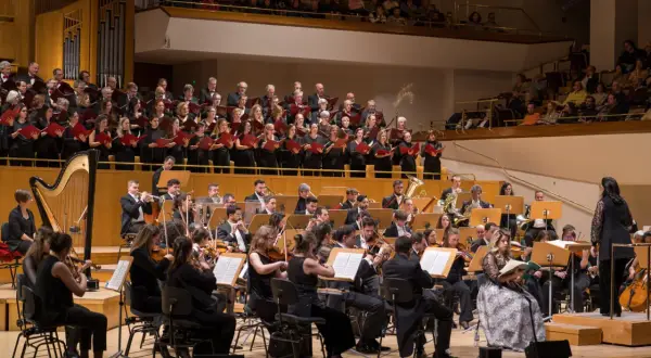 Un coro y orquesta interpretan música en un auditorio. Los cantantes franceses, vestidos de negro, sostienen partituras rojas.