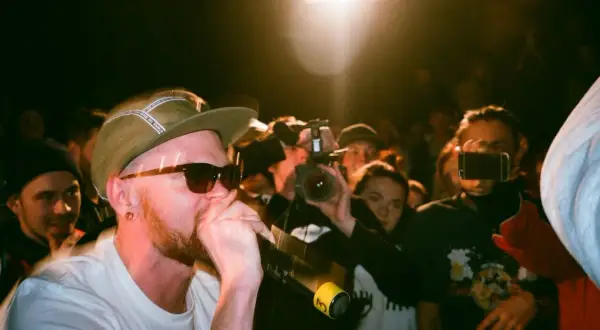 Un rapero con gorra y gafas de sol canta al micrófono, rodeado de público y cámaras. Capturando la energía de los estilos de música urbana.